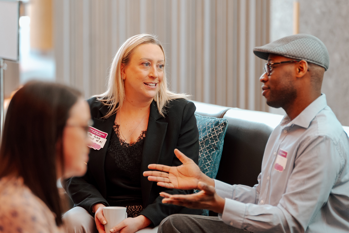 Three people sit on a couch engaged in conversation, sipping coffee. One woman smiles, while the man gestures warmly. The atmosphere is friendly.
