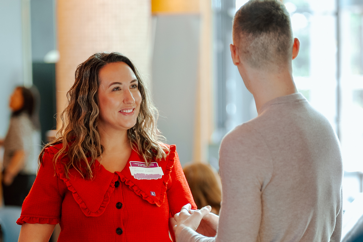 A woman in a red blouse and man in casual wear converse at a networking event. She's holding a LinkedIn-themed frame, creating a warm, professional atmosphere.