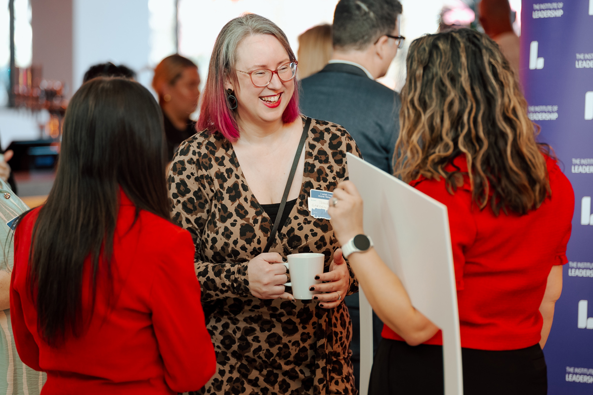 Two women in red, facing away, engage in a lively conversation with a smiling woman in a leopard print dress holding a coffee cup at a networking event.