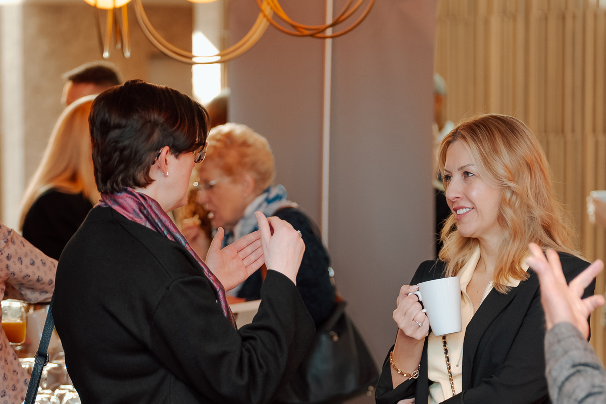 Two women smile and converse at a networking event, holding coffee cups. The atmosphere is lively, with others mingling in the background.