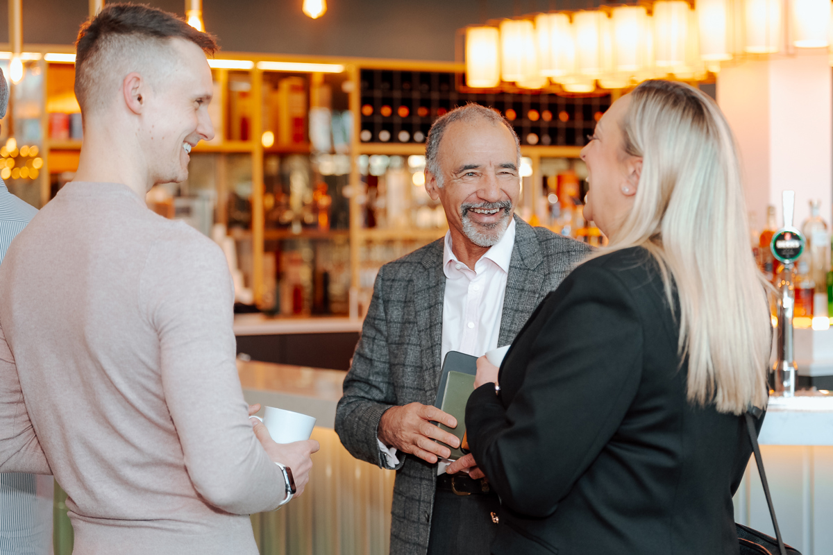 Three people smiling and talking in a warmly lit bar, one holding a phone. Shelves with bottles are in the background, creating a lively atmosphere.