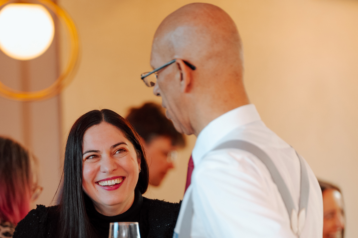 A woman and a man in formal attire share a light-hearted conversation at a social gathering, with the man holding a glass of orange juice.