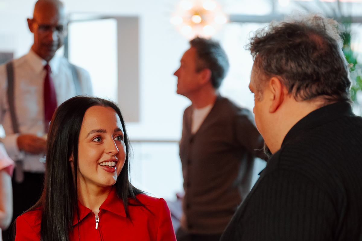 A woman in a red jacket with a name tag smiles while speaking to a man in black at an event. A pastry is on the table. The setting is lively and warm.