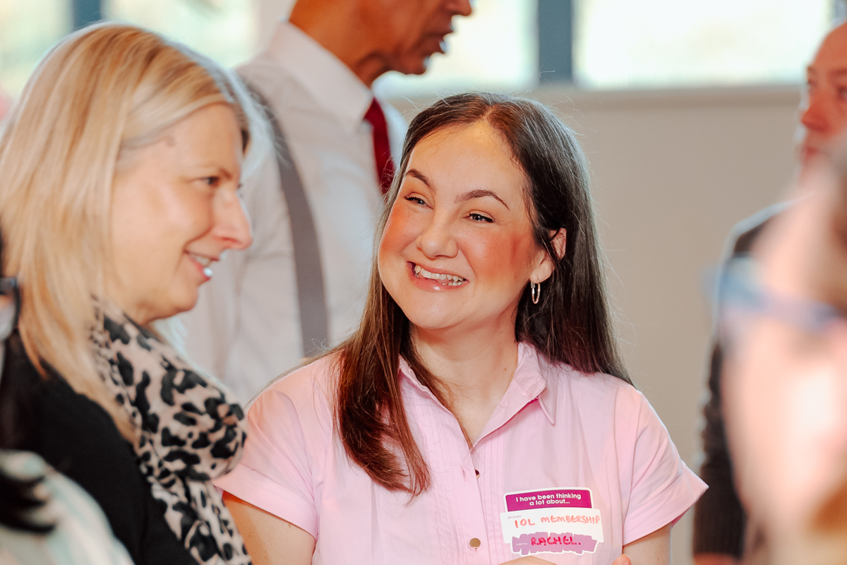 A woman in a pink dress with a name tag smiles warmly while engaging in conversation at a social gathering. The mood is lively and friendly.