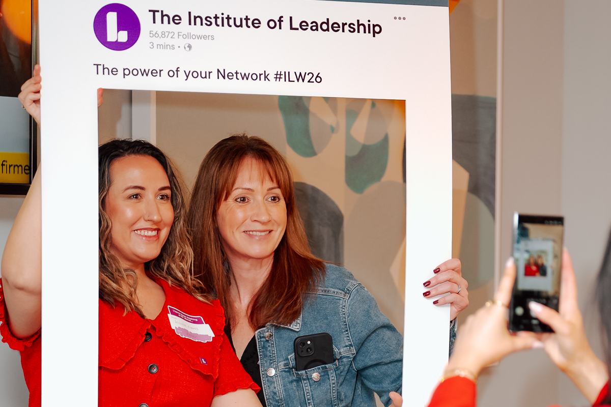 Two women smiling and posing with a large, mock LinkedIn social media frame at an event. A third person in red captures the moment on a phone.